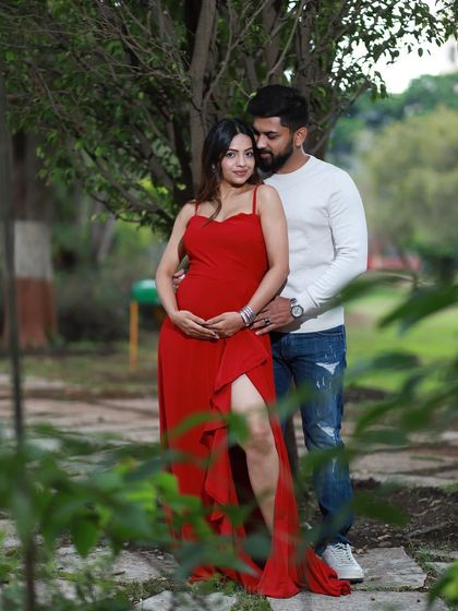 A romantic outdoor portrait surrounded by nature. The couple's connection is the focus, framed by the soft greenery and the elegance of the flowing red maternity dress.