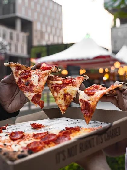 Three slices of pepperoni pizza held up against the backdrop of a food festival. It's the perfect fuel for a day of fun.