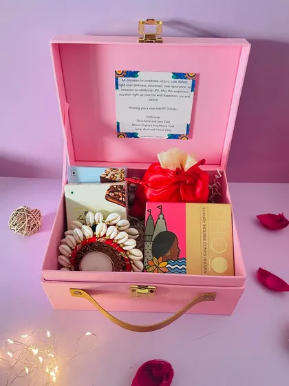 This pretty pink suitcase hamper is perfect for budget-friendly Diwali gifting. It contains shell-work T-light holders, incense, a chocolate bar, and a potli for dry fruits.