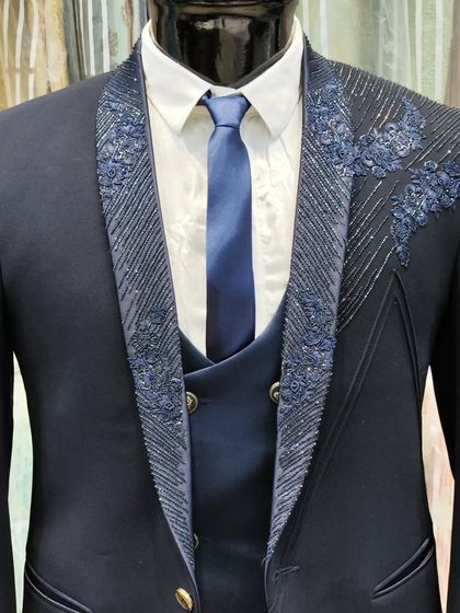 A close-up look at a hand-embroidered navy blue designer suit. The detailed beadwork and floral patterns on the lapel and shoulder showcase the high level of craftsmanship involved.