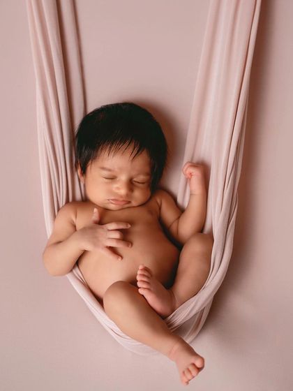 Another view of the fabric hammock pose, showing how peacefully the baby sleeps while being securely supported. The solid color background keeps the focus entirely on the baby.