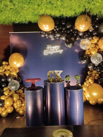 A sleek and stylish indoor birthday backdrop. This decoration uses a simple navy blue backdrop, a black and gold balloon arch, and a neon sign to create a sophisticated party corner.