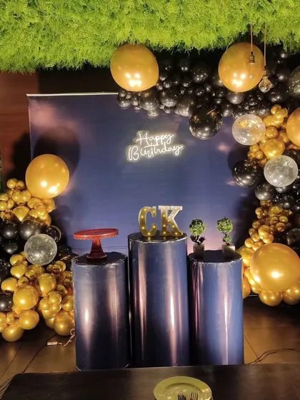 A sleek and stylish indoor birthday backdrop. This decoration uses a simple navy blue backdrop, a black and gold balloon arch, and a neon sign to create a sophisticated party corner.