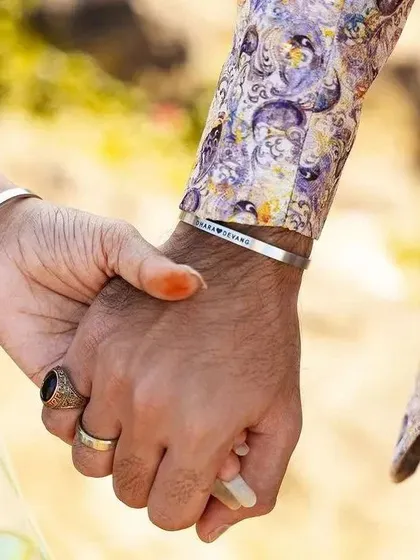 Another view of a couple holding hands, wearing matching silver cuffs engraved with their names. The man's traditional attire shows how these bracelets can be styled for special occasions.