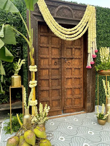 An antique-style wooden door serves as a grand entrance element, decorated with thick floral garlands and traditional brass pots.