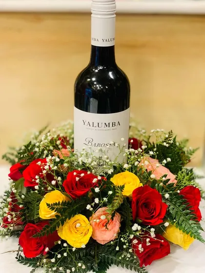 For a sophisticated and celebratory gift, I can create a beautiful floral arrangement around a bottle of wine. This example features a bottle of Yalumba wine nestled in a bed of fresh red, yellow, and pink roses with delicate baby's breath.