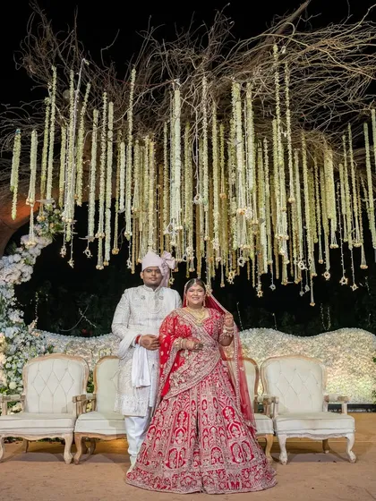 The couple poses for a portrait at their reception stage, set against a tree-of-life backdrop with hanging floral garlands. This setup provides a beautiful, serene setting for post-ceremony photographs.