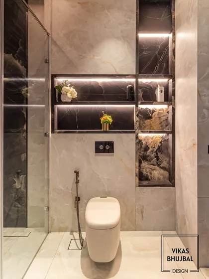 A close-up of the toilet area, highlighting the contrast between the light marble walls and the dark marble used within the storage niches. The ambient lighting adds a warm, inviting glow.