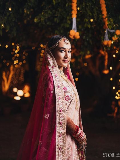 A beautiful bridal portrait taken at night. The bride, in her elegant lehenga and dupatta, is softly illuminated against a background of warm bokeh lights, creating a dreamy and romantic feel.