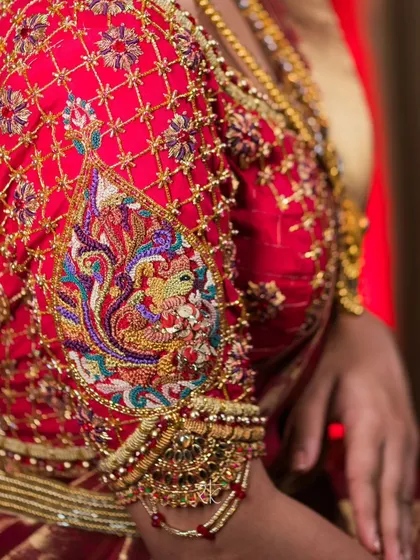 A close-up of the sleeve of our maroon bridal blouse, highlighting the complex zardosi and threadwork. The peacock motif is brought to life with a vibrant palette, set against a delicate net-like pattern of golden embroidery.
