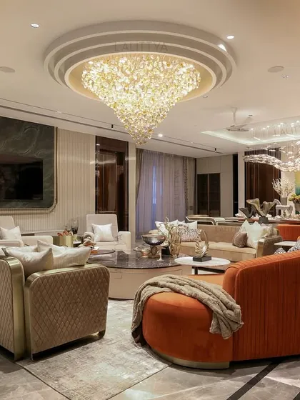 A sweeping view of a grand living room, where a curved orange sofa adds a bold splash of color. The space is anchored by a massive, tiered crystal chandelier, reflecting our love for creating dramatic and celebratory atmospheres.