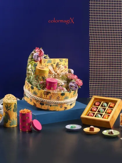 A round, tiered hamper in the yellow forest-print theme, artfully decorated with flowers and filled with gourmet jars and a box of chocolates.