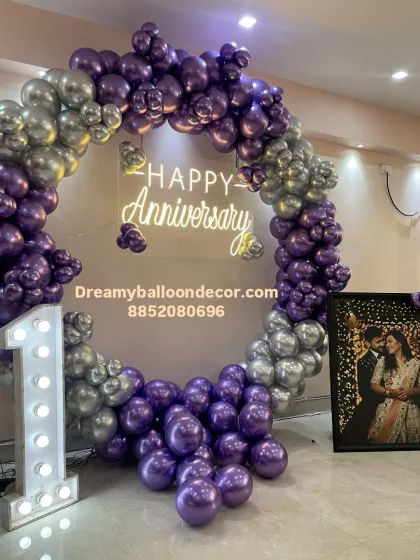 I help you celebrate your milestones in style. This first-anniversary decoration features a stunning ring backdrop with metallic purple and silver balloons, a custom 'Happy Anniversary' neon sign, and a large marquee number '1' for that perfect picture.