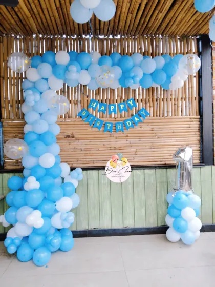 A simple blue and white L-shaped balloon arch for a first birthday against a bamboo wall.