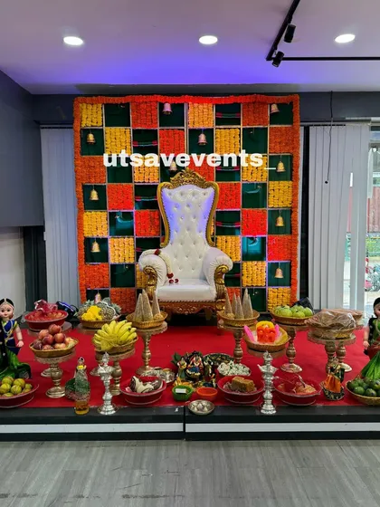 This image captures a complete and abundant seemantham plate arrangement. The stage features a multi-colored floral checkerboard backdrop, with numerous plates filled with fresh fruits, sweets, and other offerings placed on silver and gold-look stands.