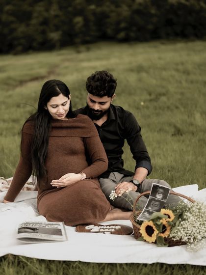 A beautiful, full-scene shot of the couple enjoying their maternity picnic in a field. They are looking down at the baby bump together, lost in their own world.