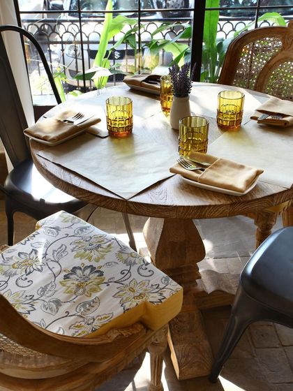 A close-up of a table setting at Amour Bistro shows the attention to detail. The rustic wooden table, patterned seat cushion, and colorful glassware all contribute to the restaurant's warm and harmonious blend of tradition and modernity.
