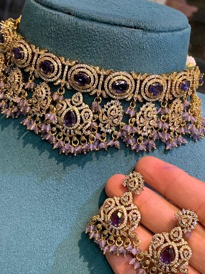 This choker set is available in multiple colors including ruby, emerald, and grey. The intricate metalwork combined with colored stones and pearl or bead droppings offers a sophisticated and royal look.