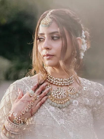A close-up portrait that captures the soft, romantic feel of this bridal look. The makeup features gentle contouring and a shimmery, neutral eye, complementing her intricate pastel and gold jewelry.