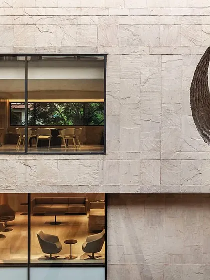 A view of the Dholpur stone façade from the courtyard, where a prominent sculpture by Subodh Gupta creates a focal point. The windows offer glimpses into the renovated office interiors.