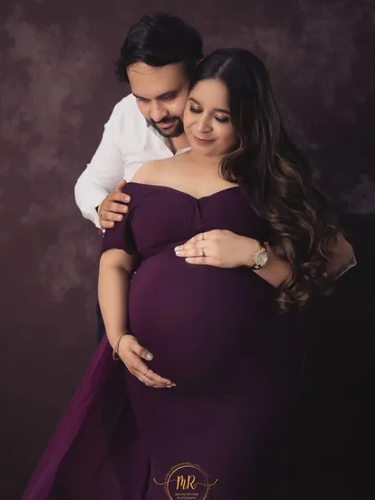 A classic studio portrait of an expecting couple. The partner stands behind his wife, both cradling the baby bump in a timeless and loving pose.