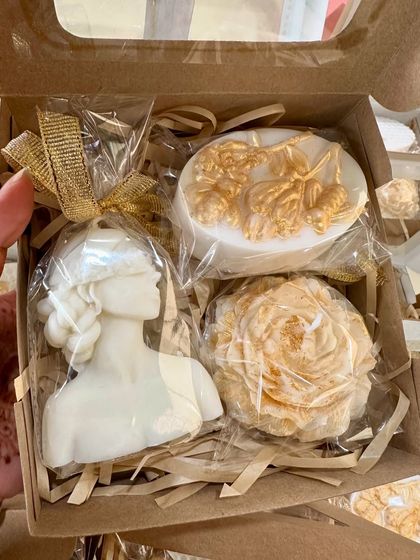 A hand holding one of the finished white and gold gift boxes. This set includes the 'Lady' candle, a peony candle, and a beautifully detailed floral soap.