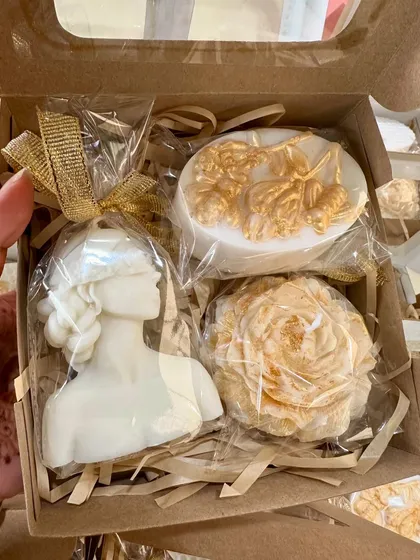 A detailed shot of the white and gold gift box, featuring a lady bust candle, a peony candle, and another ornate soap.