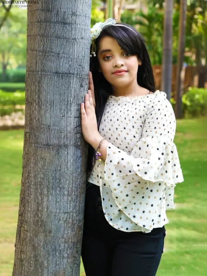 A young girl leans against a tree in a park, her polka-dot shirt and confident pose making for a stylish and natural portrait.