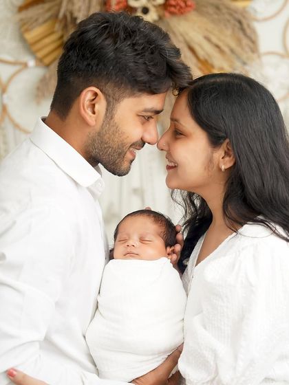 An intimate and loving family portrait. The parents' foreheads are touching as they look down at their sleeping newborn, creating a beautiful moment of connection.