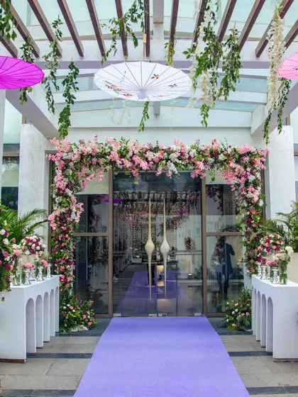 The entrance to the engagement venue, featuring a beautiful floral arch of pink and white blossoms over glass doors, leading guests down a purple carpet.