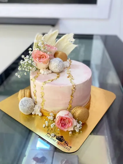 A beautiful pink and white cake decorated with fresh roses, baby's breath, macarons, and sparkling truffles. The gold chain detail adds a touch of glamour.