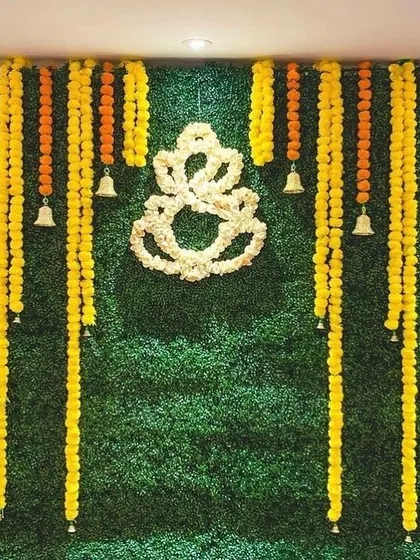 A popular choice for home festivals, this backdrop uses a green wall, marigold garlands, and a floral Ganesha outline for a traditional yet clean look.