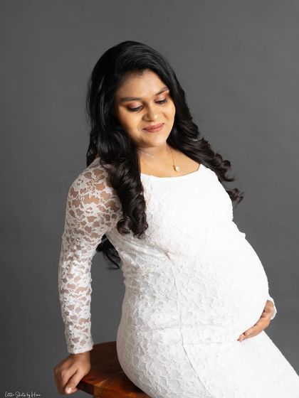 A serene portrait of a mother-to-be in a simple white lace dress. She is seated and looks down at her bump with a gentle expression, capturing a quiet moment of connection.