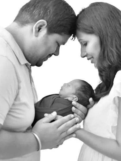 A classic black and white portrait showing the intimate connection between new parents as they hold their baby together. The focus is entirely on their loving expressions and gentle touch.