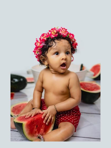 A baby girl with a surprised expression sits among watermelon slices, wearing a cute floral headband. This shot is full of personality and charm.