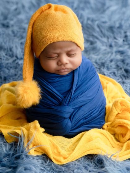 A newborn wearing a yellow pom-pom hat is swaddled in a royal blue wrap, creating a vibrant and colorful portrait.