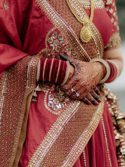 A close-up detail shot of the bride's hands, adorned with henna and red bangles, resting on her intricately embroidered lehenga.
