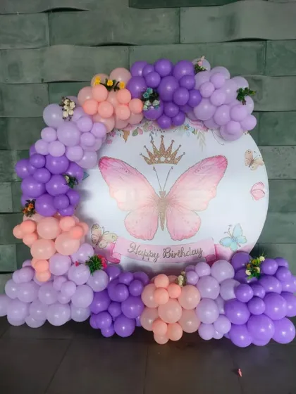 A simple and sweet butterfly birthday backdrop. A circular panel with a crowned butterfly is surrounded by a lovely balloon garland in shades of purple and peach.