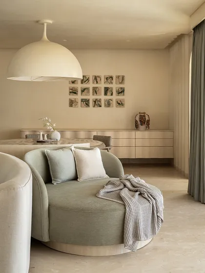 A cozy corner in the 'Soft Serve' apartment, featuring a round dining table under a large dome light. The space is designed with soft curves and a neutral palette to evoke a sense of calm and tranquility.