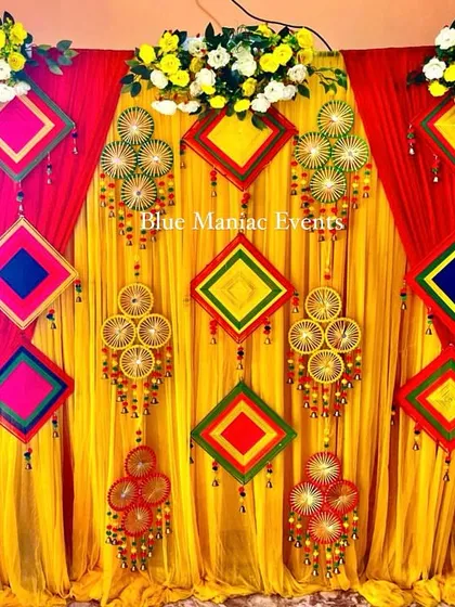A vibrant Haldi and Mehendi backdrop with red and yellow drapes and colorful traditional hangings.