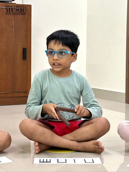 A young boy with his claves, ready to play the rhythm shown on the flash card. We use a variety of percussion instruments to keep the classes exciting and sonically diverse.