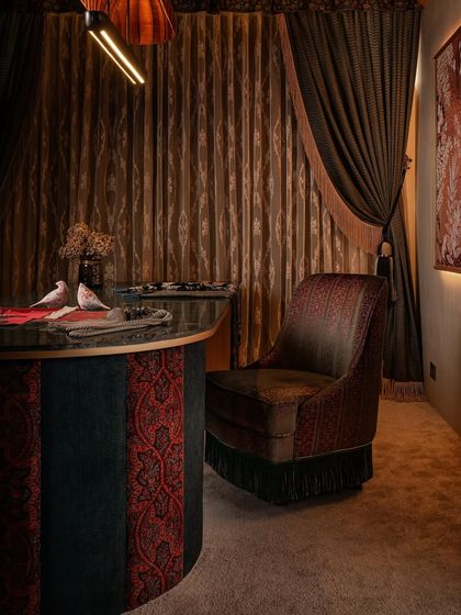 The custom-designed head chair for the client table, a swivel piece upholstered in detailed embroidery and finished with a fringe. It sits against a backdrop of heavy, layered drapes that enhance the room's maximalist and opulent feel.
