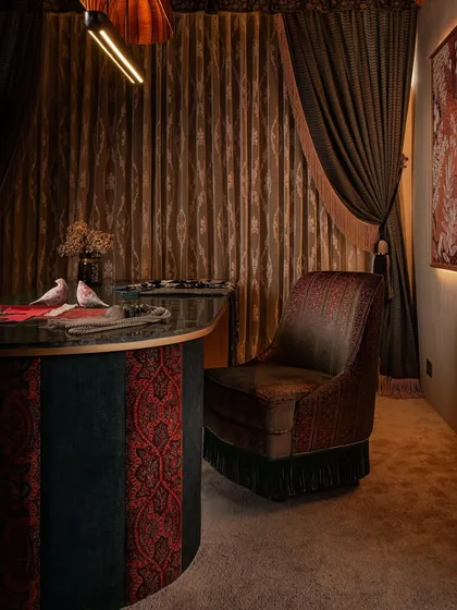 The custom-designed head chair for the client table, a swivel piece upholstered in detailed embroidery and finished with a fringe. It sits against a backdrop of heavy, layered drapes that enhance the room's maximalist and opulent feel.
