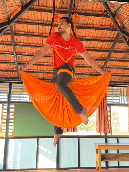 A student in an orange hammock holds the skirt pose, demonstrating the strength and balance required to hold the body stable in the air.