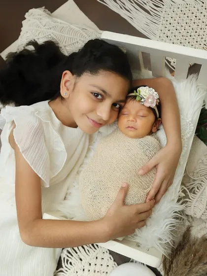 An older sister lovingly watches over her newborn sibling, who is resting peacefully in a miniature white crib. This setup is perfect for capturing the protective bond between siblings.