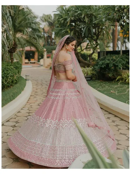 A full-length shot of the bride in her pink lehenga, showcasing the beautiful off-the-shoulder design and intricate details. The garden path setting adds a touch of natural elegance.