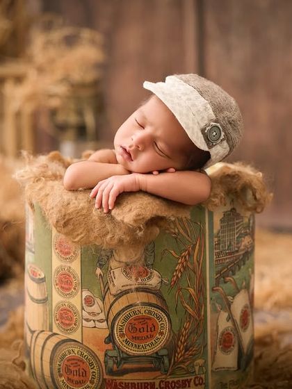 A baby wearing a vintage-style cap sleeps in a decorative tin box, surrounded by hay and other rustic props for a nostalgic feel.