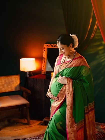 A quiet, contemplative portrait of a mom-to-be in a green saree. The soft, warm light from the lamp creates an intimate and peaceful atmosphere.