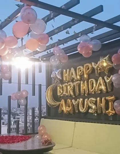 A romantic rooftop birthday surprise for Aayushi. The setup includes 'Happy Birthday' foil letters, pastel balloons, and a table with scattered rose petals.