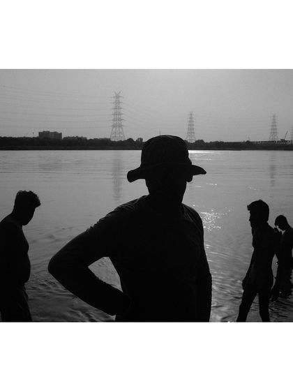 A black and white silhouette of several people wading in the Yamuna River. The strong backlighting from the sun creates a high-contrast, graphic image.